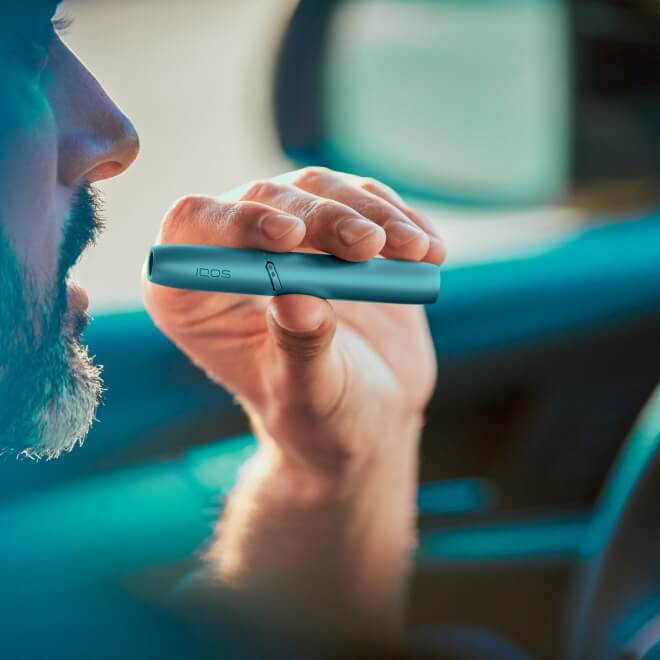 Hand holding a turquoise IQOS Originals One heated tobacco device with a guitar.